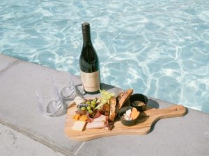 Wooden board with meats and cheeses, and bottle of pinot noir sitting next to edge of pool