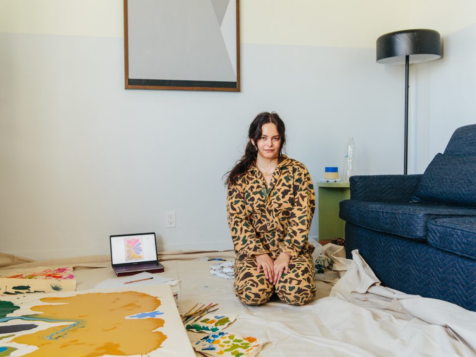 Woman wearing camo pajamas, keeling on drop cloth, next to large canvas and open laptop with flower sketch