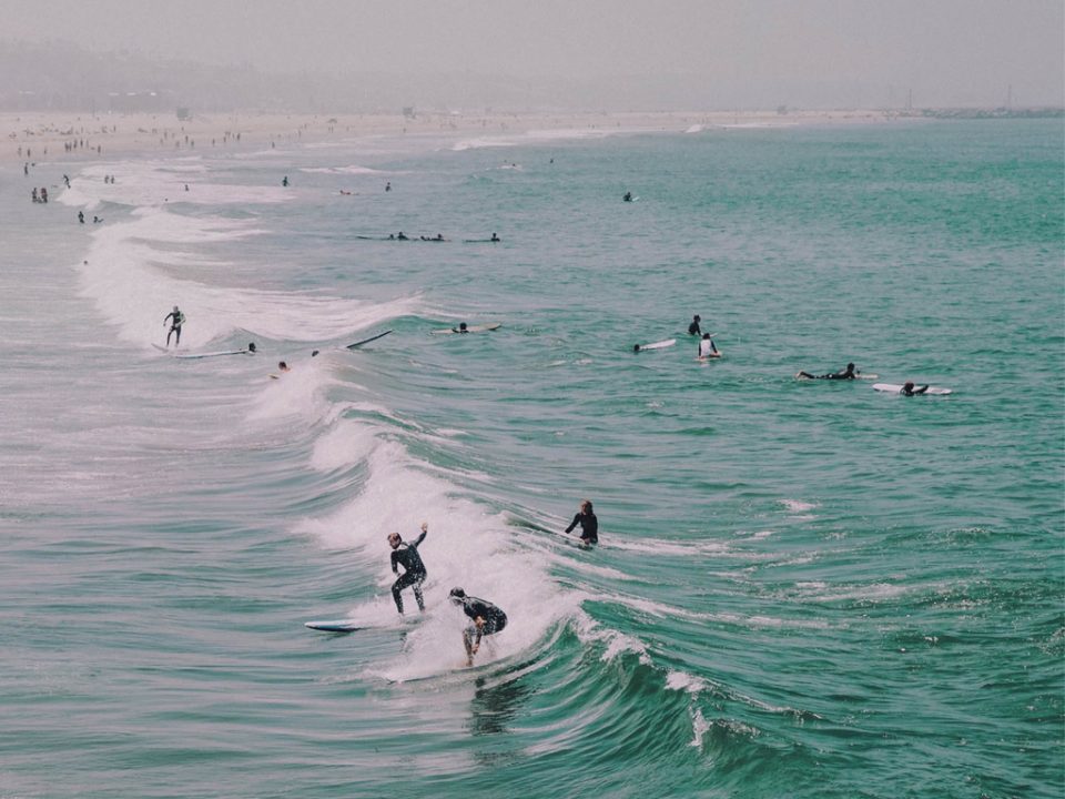Surfers catching waves along busy beach coastline with multiple people in the water