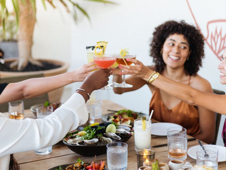 Women smiling, toasting with cocktails, sitting around a wooden table filled with entrees and drinks