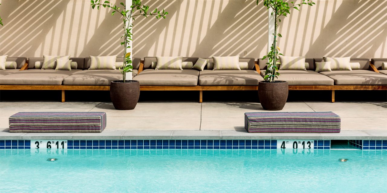 Pool in the foreground, padded cushion next to edge, potted plants and lounge chairs under cabanas