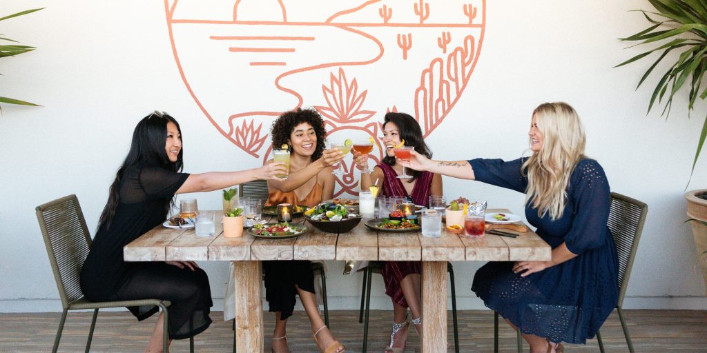 Women smiling, toasting with cocktails, sitting around a wooden table filled with entrees and drinks