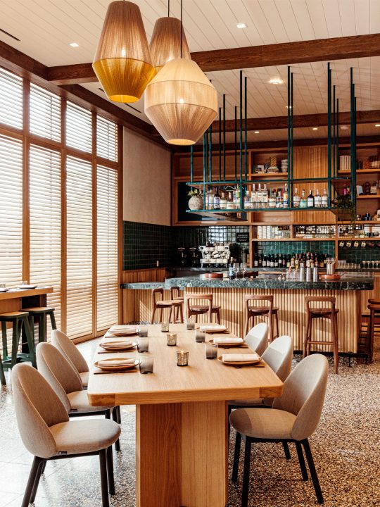 Wooden restaurant table, chair, lamps overhead, and bar with stool chairs in the background